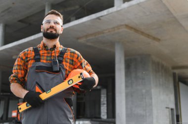 İnşaat işçisi inşaat alanında duruyor, yeni projeler için sağlam bir şapka ve seviye tutuyor.