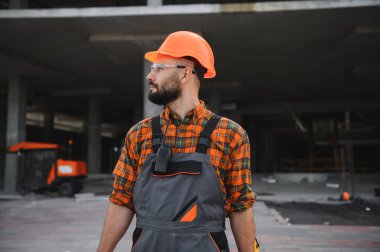 Kasket ve güvenlik gözlüğü takan erkek inşaat işçisi gelişmekte olan bir inşaat alanına bakıyor.