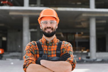 Sağlam şapka ve güvenlik gözlüğü takan kendine güvenen inşaat işçisi inşaat alanında çapraz kollarla dururken gülümsüyor.