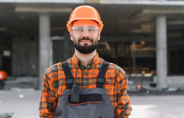 İnşaat işçisi inşaat sahasında duruyor. Koruyucu iş kıyafeti, güvenlik gözlüğü ve şapka takıyor.