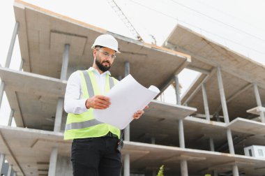 Güvenlik yeleği ve şapka takan mühendis planları inceliyor, inşaat halindeki bir binaya karşı duruyor.