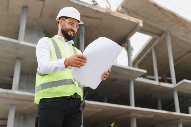 Gülümseyen inşaat mühendisi. Çağdaş bir inşaat alanındaki inşaat planlarını gözden geçiriyorlar.