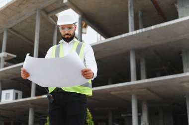 Modern bir inşaat sahasının planlarını inceleyen sert şapka ve güvenlik yeleği giymiş profesyonel erkek mühendis ya da mimar.