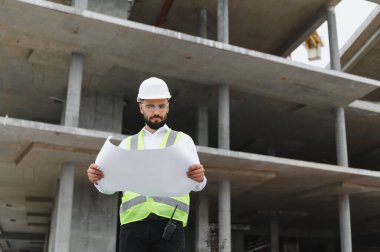 Sert şapka ve güvenlik yeleği giyen profesyonel bir adam beton inşaat alanındaki bina planlarını inceliyor.
