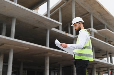 Çelik yelek ve şapka giyen erkek mühendis modern bir inşaat alanının planlarını inceliyor.