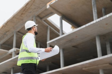 İnşaat mühendisi proje planlarını tutuyor, modern bir inşaat sahasının inşaatını denetliyor.