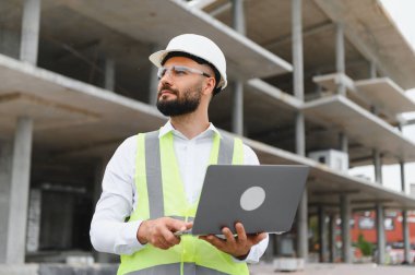 Sert şapka ve güvenlik yeleği giyen mühendis, beton inşaat alanındaki planlara bakıyor.
