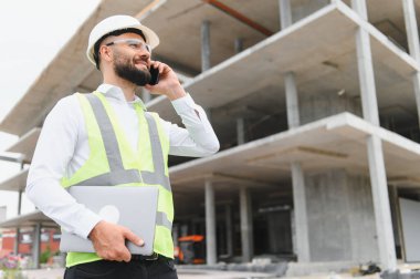 Gülümseyen inşaat mühendisi, çelik yelek giyiyor, dizüstü bilgisayar tutuyor, iskeletin önünde telefonla konuşuyor.