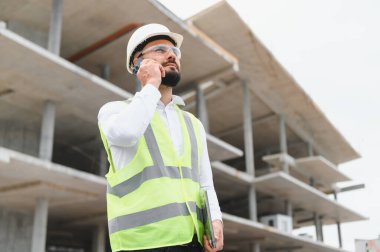 İnşaat alanında inşaat başlığı ve güvenlik yeleği mühendisi akıllı telefondan konuşuyor, ilerlemeyi denetliyor ve proje planlamasını yapıyor.
