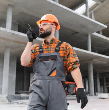 Sakallı, kasketli, güvenlik gözlüklü ve tulumlu bir işçi bitmemiş bir binada telsize konuşuyor.