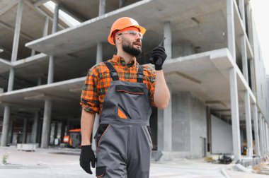 İnşaat işçisi, başlık ve güvenlik gözlüğü takıyor, inşaat alanında çalışırken telsizle iletişim kuruyor.