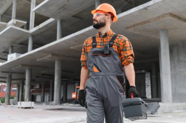 İnşaat işçisi, kasketli, güvenlik gözlüklü ve tulumlu. Bitmemiş beton bir inşaat sahasında duruyor. Elinde alet çantası var.