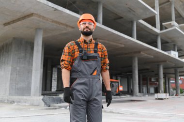 Güvenilir inşaat işçisi, sağlam bir şapka, güvenlik gözlüğü ve tulum takıyor, bitmemiş beton bir binanın önünde duruyor.