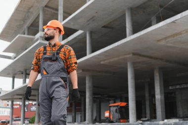 Başlık, tulum ve güvenlik gözlüğü takan genç bir müteahhit, gururla duruyor, gelişimi denetliyor.