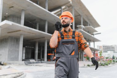 Tecrübeli inşaat işçisi ya da mühendis güvenlik ekipmanı giyiyor ve inşaat alanında telsizle iletişim kuruyor.