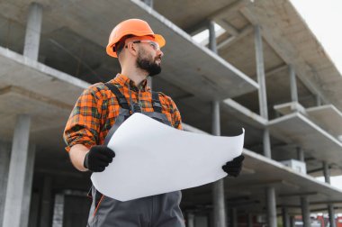 İnşaat alanında güvenlik başlığı ve gözlük takan mühendis ya da mimar gelişimi denetliyor.