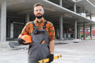 Güvenilir bir inşaat işçisi, güvenlik gözlüğü takıyor, sıkı bir şapka takıyor ve inşaat alanında duruyor.