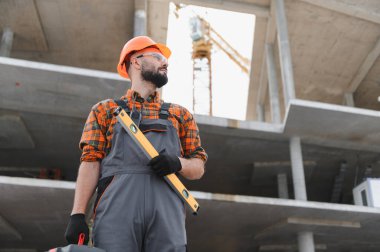 Başlık ve güvenlik gözlüğü takan inşaat işçisi elinde bir aletle inşaat sürecini bir şantiyede inceliyor.