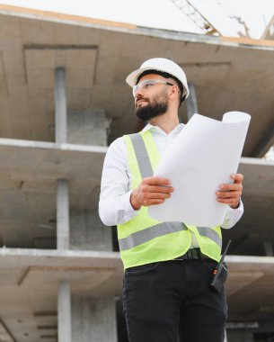 İnşaat alanındaki güvenlik planlarını gözden geçiren sert şapka ve çelik yelek giyen profesyonel erkek mühendis.
