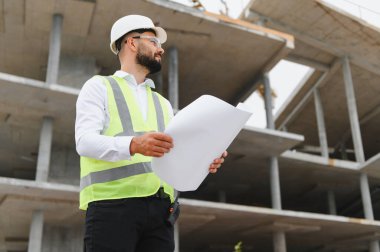 Bir inşaat alanının planlarını inceleyen, proje geliştirme çalışmalarını denetleyen, çelik yelekli bir adam.
