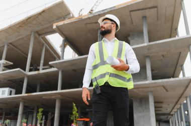 Genç bir mimar ya da inşaat işçisi sıkı bir şapka ve güvenlik yeleği giyer, inşaat alanında dikilirken gözlerini kaçırır.