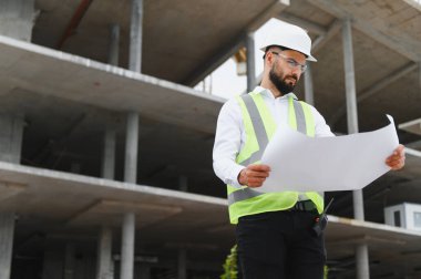 İnşaat alanındaki inşaat planlarını inceleyen mühendis koruyucu giysiler giyerken projenin ilerlemesini ve güvenliğini sağlıyor.