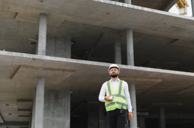 Mühendis ya da mimar, çelik yelek ve şapka giyen, elinde planlar tutan, inşaat alanını inceleyen.