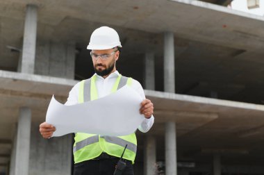 Güvenlik miğferi ve yeleği olan mühendis inşaat planlarını gözden geçirip proje geliştirmeyi planlıyor.