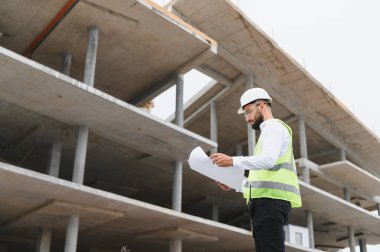 Güvenlik kaskı ve yelek giyen mühendis tamamlanmamış beton bir yapının planlarını inceliyor.