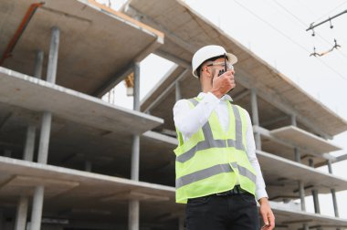 İnşaat işçisi, çelik yelek ve şapka giyiyor. İnşaat alanında telsizle iletişim kuruyor.