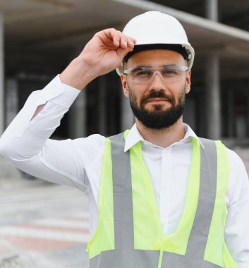 Kendine güvenen sakallı mühendis ya da mimar çelik yelek giyip inşaat alanındaki kameraya bakıyor.