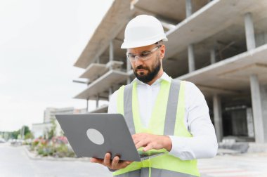 Erkek inşaat mühendisi sert bir şapka ve güvenlik yeleği giyiyor, dışarıda bir inşaat alanında dizüstü bilgisayar kullanıyor.