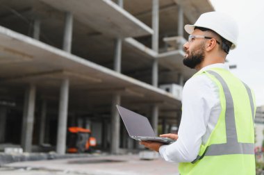 Güvenlik kaskı ve yeleği giyen profesyonel mühendis dizüstü bilgisayarı tutuyor, inşaat projesini denetliyor.