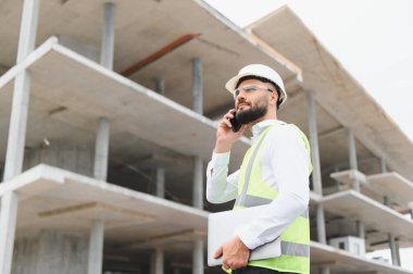 Erkek mühendis, çelik yelek ve şapka giyiyor, dizüstü bilgisayar tutuyor ve inşaat halindeki bir binada telefonla konuşuyor.