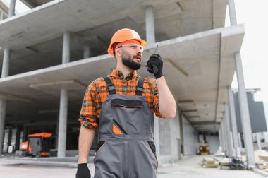 İnşaat işçisi, inşaat başlığı ve güvenlik gözlüğü takıyor, bitmemiş beton bir binanın önünde dikilirken telsiz kullanıyor.