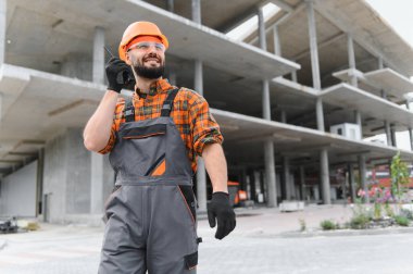 İnşaat işçisi, başlık ve güvenlik gözlüğü takıyor, telsizle iletişim kuruyor, gülümsüyor, inşaat alanında duruyor.