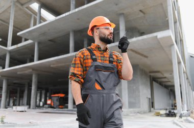 İnşaat işçisi, başlık ve güvenlik gözlüğü takıyor, aktif bir inşaat alanında telsizle iletişim kuruyor.