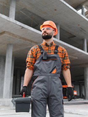 Başlık ve güvenlik gözlüğü takan mühendis ya da ustabaşı, inşaat alanında bir alet çantası taşıyor.