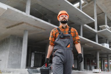 Sert şapkalı ve tulumlu bir adam bitmemiş bir inşaat alanında alet çantası taşıyor.