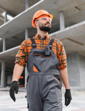 İnşaat işçisi kişisel koruma ekipmanlarıyla inşaat alanında duruyor, kendinden emin bir şekilde ileriye bakıyor.