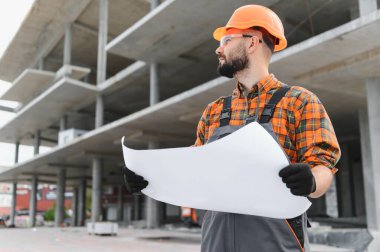 Güvenlik ekipmanı takan bir mühendis, elinde bir plan tutuyor ve bina yapısını geliştiriyor. İnşaatı planlama ve denetleme