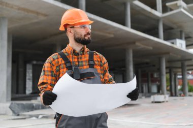 Güvenlik kasketi ve gözlük takan sakallı adam inşaat alanında çalışırken elinde planlar tutuyor.