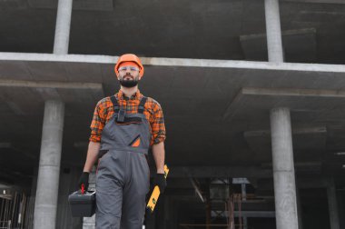 İnşaatçı sabit şapka ve güvenlik gözlüğü takıyor. Alet çantası taşıyor ve beton bir inşaat sahasında duruyor.