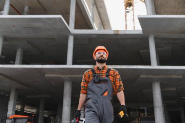 İnşaat işçisi baret ve güvenlik gözlükleriyle beton bir inşaat sahasında duruyor, aletleri tutuyor, ileriye bakıyor.