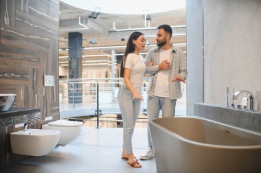 Genç çift seçenekleri tartışıyor ve modern bir banyo galerisinde çeşitli eşyalar eşliğinde ev dekorasyonu planlıyor.