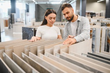 Modern bir sergi salonundaki döşeme örneklerini karşılaştıran genç bir çift, ev yenileme ve iç tasarım malzemelerine karar veriyorlar.