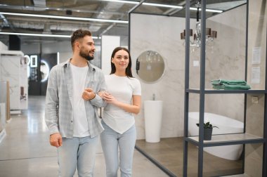 Banyo yenileme fikirleri arayan genç bir çift modern bir ev geliştirme galerisinde kol kola yürüyorlar.