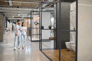 Genç çift kol kola modern bir galeride banyo kiremit örneklerini inceliyor, bir ev yenileme ve tasarım güncellemesi planlıyor.