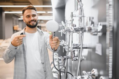 Genç adam banyo malzemeleri alıyor, duş başlığı tutuyor ve bir nalbur dükkanında bir dizi seçeneğe işaret ediyor.