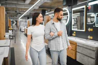 Ev dekorasyonu mağazasında yürüyen, banyo mobilyalarına ve aynalara bakan, evi yenilemeyi planlayan bir çift.